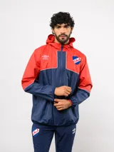 Campera de lluvia Umbro del Club Nacional de Football, con diseño de bloques de color en rojo y azul marino. Presenta capucha, cierre frontal, puños elásticos y detalles de piping blanco. Incluye logo de Umbro y escudo del club en el pecho.