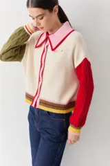 Cardigan de punto con diseño color block, cuerpo en color crema, cuello polo rosado con borde rojo, manga derecha verde oliva y manga izquierda roja. Presenta mangas con tejido de ochos, puños acanalados multicolor y cierre frontal con botones.