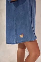 Vestido corto de denim azul, corte camisero holgado, mangas cortas amplias, cuello clásico, cierre frontal con botones, bolsillo aplicado en el pecho y pequeño bordado lateral en el dobladillo.