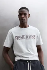 Camiseta de algodón gris melange con cuello redondo, mangas cortas y efecto lavado. Presenta un estampado de texto frontal en color oscuro con efecto desgastado.