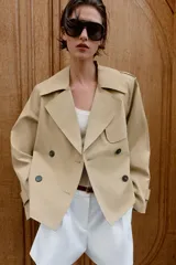 Trench corto de color beige, con cuello de solapa, manga larga con hombreras y trabillas con botones. Cuenta con cierre frontal cruzado con botones oscuros y cinturón ajustable del mismo tejido.