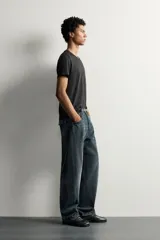 Jeans loose fit de hombre en denim de algodón color gris oscuro con efecto lavado. Presenta perneras con costuras giradas hacia los tobillos, tiro medio, cierre frontal con botones y bolsillos frontales con bolsillo cerillero.