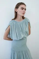 Vestido midi de tejido texturizado color verde menta, con escote redondo fruncido, espalda semiabierta con breteles finos y capa decorativa sobre los hombros. Cuenta con sección de punto smock en la cadera y falda con leve vuelo.
