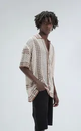 Camisa de manga corta tejida en punto abierto (símil crochet), color beige con detalles en marrón. Presenta cuello solapa, cierre frontal con botones y calce oversize.