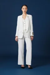 Saco de mujer color blanco, con solapas clásicas, cierre frontal de un botón y bolsillos laterales con solapa. Presenta un diseño estructurado con un parche decorativo en la manga izquierda.