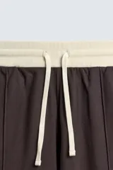 Pantalón jogger de corte wide fit en tejido compacto color gris oscuro. Presenta cintura elástica con cordón ajustable, bandas laterales en contraste color beige y costuras frontales tipo pinza.