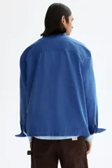 Sobrecamisa de lona de algodón en color azul, con cuello clásico, cierre frontal de botones y tres bolsillos de parche en la parte delantera. Presenta mangas largas con puños abotonados y canesú en la espalda.