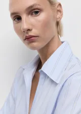 Camisa de corte recto con diseño de rayas verticales en tonos celestes y blancos. Presenta cuello camisero con solapas amplias, cierre frontal de botones ocultos bajo tapeta y mangas largas con puños abotonados.