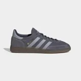 Championes Adidas Handball Spezial de gamuza color gris con las icónicas tres tiras laterales en un tono más claro y suela de caucho natural. Presenta el logo de la marca y el nombre del modelo en dorado en el lateral.