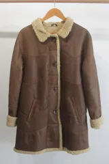 Campera tipo gamulán de cuero genuino en color marrón, con interior y detalles en corderito sintético color crema. Presenta cierre frontal con botones, cuello solapa y puños con vuelta.