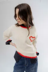 Buzo de punto color crema con detalles en rojo en el cuello, puños y ruedo. Presenta un bolsillo frontal en el pecho con un corazón bordado en relieve.