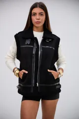Chaleco negro de estilo aviador con interior y bordes de corderito sintético. Presenta cierre frontal metálico, detalles de tiras de cuero sintético y hebillas ajustables en el cuello y el ruedo.