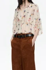Camisa oversize de tejido fantasía color crema con estampado de pájaros y flores en tonos azules, amarillos, naranjas y rojos. Presenta finas rayas verticales en colores pastel y lentejuelas distribuidas por toda la prenda. Tiene cuello camisero, cierre frontal con botones y mangas cortas rectas con un ligero vuelo. Incluye un bolsillo de plastrón en el pecho izquierdo.