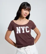 Remera de algodón color marrón oscuro con escote amplio tipo barco u hombro a hombro y manga corta. Presenta el estampado en mayúsculas "NYC" en color blanco en el frente.