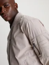 Campera tipo bomber de Calvin Klein en color beige, confeccionada en una mezcla de algodón y poliamida. Presenta cuello alzado, puños y bajo de punto canalé, cierre frontal de cremallera, bolsillos laterales de hendidura y un bolsillo adicional con cremallera en la manga izquierda con insignia de goma de la marca.