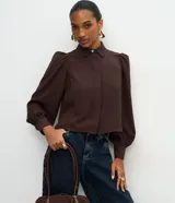 Camisa de crepé color celeste, con cuello inglés, manga larga abullonada y cierre frontal oculto con botón diferenciado en el cuello.