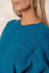 Buzo de punto color azul petróleo, con cuello redondo, mangas largas con puños ajustados y detalles de textura trenzada en la parte superior y el ruedo.