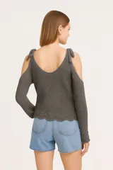 Sweater de tejido de punto color natural, con diseño de hombros descubiertos, mangas largas y detalle de lazos anudados en los hombros. Presenta un cuello en V profundo y un borde inferior con terminación ondulada.