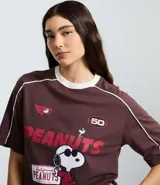 Remera de algodón color marrón con cuello redondo y mangas cortas. Presenta un estampado frontal con el personaje Snoopy en temática de carreras y detalles de líneas blancas contrastantes en los hombros y mangas.