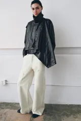 Chaqueta de efecto piel en color negro con cuello subido y bufanda desmontable integrada mediante botones. Presenta manga larga con trabillas ajustables en los puños, cierre frontal con cremallera y bolsillos laterales ocultos en las costuras. El diseño incluye un bajo con elástico en la espalda y forro interior.