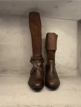Bota de montar de caña alta, confeccionada en cuero marrón con combinación de texturas. Presenta un diseño con puntera redondeada, taco cuadrado y una correa decorativa cruzada con tachas metálicas en el tobillo.