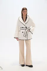 Chaqueta blanca de corte holgado con capucha, cierre frontal cruzado con botones y cinturón de tela ajustable. Presenta detalles de costuras decorativas en color negro en los bordes, solapas, bolsillos y cinturón.