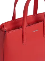 Cartera tote pequeña de piel sintética reciclada color rojo, con asas de mano fijas y correa ajustable opcional. Presenta cierre de cremallera superior, bolsillos interiores con ranura y logo metálico de Calvin Klein en el frente.