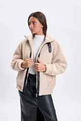 Campera de sherpa color crudo con detalles en cuero sintético negro en el cuello y la solapa frontal. Cuenta con cierre frontal y cuello alto con hebilla ajustable.