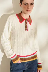 Buzo tejido de punto color marfil, con diseño de trenzas, cuello polo marrón con borde fucsia y cierre frontal parcial con botones. Presenta detalles de rayas multicolor en los puños y el ruedo, y un pequeño bordado en el pecho.
