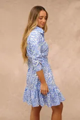 Vestido corto camisero con estampado animal print en tonos celeste y blanco, de manga tres cuartos, con cierre frontal de botones, lazo ajustable en la cintura y detalle de volados en el ruedo.