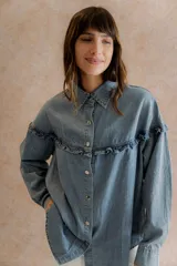 Camisa de denim liviano con calce relajado, cuello camisero clásico y cierre frontal con broches metálicos. Presenta un canesú con volado en el delantero y la espalda, mangas largas con puños abotonados y terminación inferior redondeada.