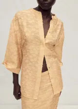 Camisa oversize de color beige con cuello de tira y cierre frontal de botones ocultos. Presenta mangas tres cuartos y un tejido con textura irregular.