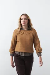 Sweater de tejido suave color tostado, con cuello redondo y detalle de flores bordadas en relieve en la parte superior del pecho. Presenta un corte relajado y mangas largas con puños ajustados.