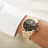 Reloj Michael Kors modelo Bradshaw, con caja y pulsera de acero inoxidable dorado, esfera negra con números romanos y tres subesferas de cronógrafo.
