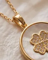 Collar de bronce dorado con cadena fina y dije redondo que presenta un fondo de nácar blanco y un trébol de cuatro hojas en el centro.