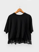 Remera negra de corte holgado y diseño minimalista, confeccionada en algodón. Presenta cuello redondo, mangas cortas y un delicado volado de encaje en el ruedo inferior.