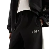 Pantalón New Balance modelo Trackside Woven, color negro con detalles laterales en gris y logo reflectante en el muslo. Cuenta con cintura elástica y puños elastizados en los tobillos.