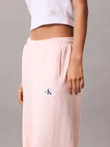 Pantalón de felpa francesa rosa pastel, de corte holgado con basta ancha y tiro medio. Posee cintura elástica, bolsillos laterales y una pequeña insignia blanca con el logotipo de Calvin Klein en el lateral izquierdo.