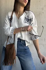 Camisa blanca de manga larga con escote en V y cierre frontal de botones. Presenta delicados bordados florales en tono oscuro en los hombros y puños.