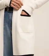 Cardigan de punto color blanco con detalles en negro en los bordes de los bolsillos, puños y ruedo. Presenta un diseño abierto, corte alargado y mangas largas.