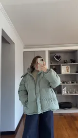 Campera puffer de corte holgado en color beige, con diseño acolchado, cuello alto y capucha desmontable. Cuenta con cierre frontal y botones a presión, además de puños elásticos.