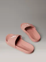 Ojotas tipo slide Calvin Klein Jeans, color rosado, con banda superior de lona de algodón reciclado y logo de la marca estampado en blanco. Presentan suela de EVA con plantilla ergonómica moldeada.