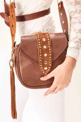 Cartera bandolera de cuero graneado color tabaco y gamuza color óxido. Presenta una tapa frontal decorada con una franja de gamuza con tachas metálicas, un colgante de flecos laterales y una correa larga ajustable. Incluye un bolsillo con cierre en la parte posterior de la tapa.