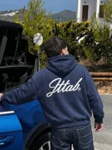 Hoodie azul marino con capucha y bolsillo frontal tipo canguro. Presenta un estampado gráfico en la espalda con la palabra 'Jttab' en tipografía cursiva blanca.