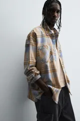 Camisa de manga larga con cuello solapa y dos bolsillos frontales de plastrón con botón. Presenta un estampado de cuadros en tonos beige, azul claro y marrón.