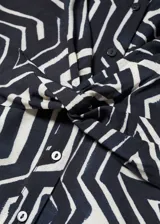 Vestido corto de corte recto y tejido fluido, con estampado geométrico en blanco y negro. Presenta cuello camisero, mangas cortas y un detalle de nudo en la cintura que realza la silueta.