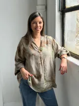 Blusa de lino color visón con estampado botánico en tonos marrones y verdes, cuello en V con abertura y un botón grande decorativo. Tiene mangas largas con tirilla para sujetar arremangadas y corte holgado.