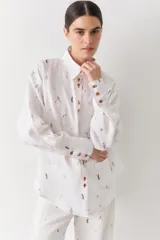 Camisa de lino blanca con estampado de figuras deportivas pequeñas y detalles de costuras en color rojo. Presenta cuello clásico, cierre frontal con botones de madera, mangas largas con puños abotonados y un bolsillo frontal aplicado.