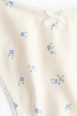 Pack de cinco bombachas tipo colaless de algodón, de tiro bajo con cintura en V, bordes con acabado picot y detalle de lazo pequeño en el centro. Incluye tres lisas en tonos azul, celeste y rosado, y dos con estampado floral pequeño sobre base clara.