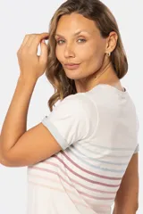 Remera de manga corta con cuello redondo, color base crema, con rayas horizontales en tonos pastel como rosado, azul claro y verde pálido. Los puños de las mangas son doblados y tienen un borde de color celeste pálido.